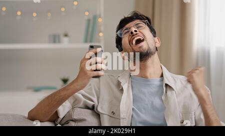 Junger 30s araber bärtiger Mann Millennial Kerl in Brille sitzt zu Hause auf der Couch Blick auf Handy macht ja Geste immer Angebot gewinnen gute Nachrichten Stockfoto