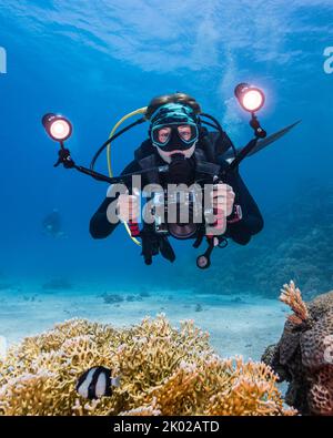 Nahaufnahme eines Tauchers mit einer Unterwasserkamera mit zwei Lichtern, die auf die Kamera mit einem Korallenriff im Vordergrund zeigen Stockfoto