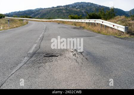 Gebrochener Fleck mitten auf einer Straße, die sich auf dem Land gegen hohe Waldberge und Hügel erstreckt Stockfoto