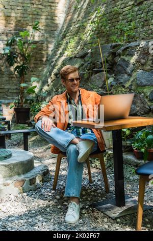 Fröhlicher Rotschopf-Mann in Sonnenbrille, der aus der Ferne auf dem Laptop arbeitet, während er auf der Außenterrasse sitzt, Stockbild Stockfoto