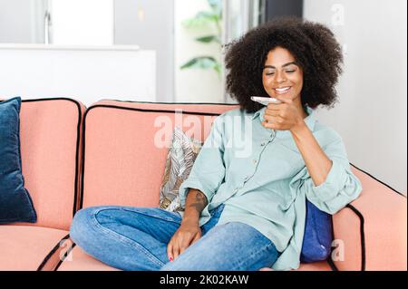 Junge afrikanerin, die Sprachnachrichten sendet und Notizen über das Smartphone aufnimmt, sitzt auf dem Sofa in einer gemütlichen, modernen Wohnung zu Hause und nutzt die Spracherkennungs-Funktion Stockfoto
