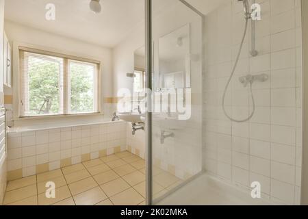 Badewanne mit Glasdusche in der Ecke neben Waschbecken und Spiegel im kleinen hellen Badezimmer mit weißen Kachelwänden Stockfoto