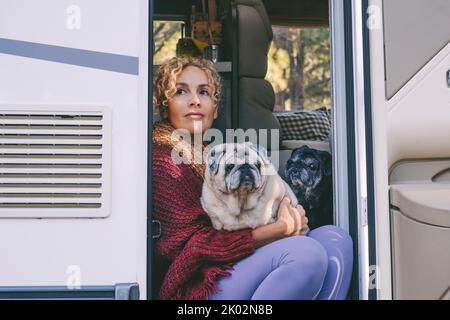 Nette Erwachsene Frau sitzt und entspannt auf der Tür ihres Wohnwagens mit ihren zwei Mops Hunde. Beste Freunde und Freundschaft Konzept. Van Leben und Reise Straße Urlaub Leben Urlaub. Moderne weibliche Menschen Stockfoto