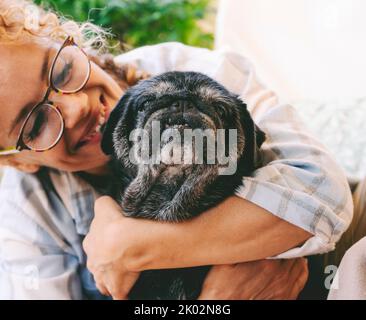 Fröhliche Frau umarmt schwarzen alten Hund mit Liebe und Freindship. Tierischer Mops bester Freund für immer. Weibliche Menschen und ihr Welpe genießen Zeit zusammen mit Glück. Lady lächelt und bindende Hund Stockfoto