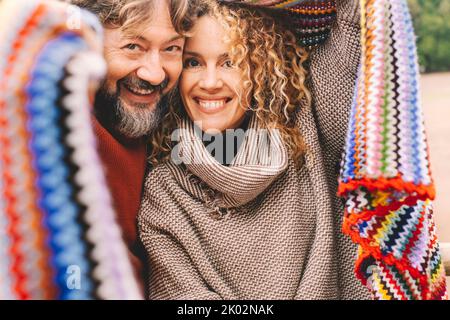 Porträt eines jungen, reifen Paares, das lächelt und gemeinsam unter einem bunten Strickmantel aus Wolle die Freizeit genießt. Liebe und Freundschaft zwischen Mann und Frau. Menschen, die Spaß haben und lachen. Urlaub Stockfoto