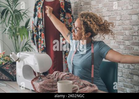 Frau streckt die Arme nach der Arbeit an der Nähmaschine zu Hause oder Design-Bekleidungsstudio. Weiblicher Stretching Körper nach Überlastung mit Kleidung Reparatur oder Produktion. Konzept der alternativen Arbeit in Innenräumen Stockfoto