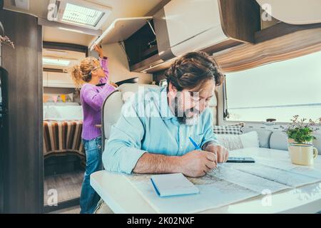 Bärtiger Mann, der den nächsten Reiseurlaub plant, sitzt am Tisch in seinem Wohnmobil. Frau im Hintergrund Kochen und Verwaltung der Lagerung. Urlaub im Urlaub Stockfoto