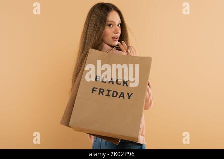 Lächelndes Teenager-Mädchen mit Papiertüten mit schwarzem freitag Schriftzug isoliert auf beige, Stockbild Stockfoto