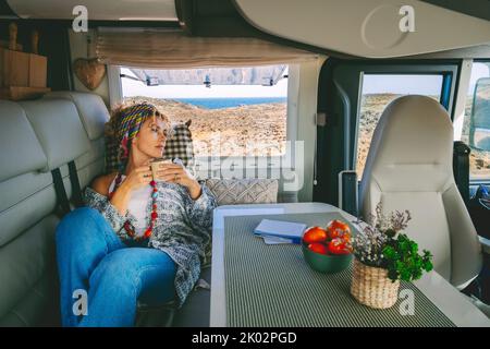 Nette trendige Erwachsene Frau haben Entspannung Freizeitbeschäftigung liegt auf dem Sofa in ihrem gemütlichen Alternative Wohnmobil. Konzept des Tourismus und moderne Vanlife. Blick auf den Strand und das Meer vor dem Fenster Stockfoto