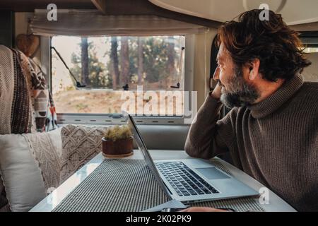 Beruflicher Lifestyle abseits der Grenzen mit einem reifen Mann, der in einem Wohnmobil an einem Laptop arbeitet und vor dem Fenster in den Wald blickt. Van Life People Konzept. Remote-Mitarbeiter, der eine Online-Verbindung für den Computer verwendet Stockfoto