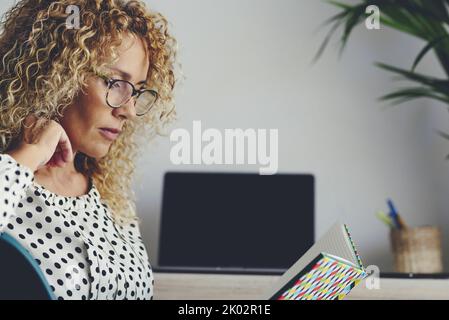 Seitenportrait einer niedlichen erwachsenen Dame, die ein Notizbuch mit Brille liest. Konzept für Schüler im Alter von Jahren zu Hause mit Laptop im Hintergrund. Moderne Single Lifestyle weibliche Menschen lesen ein Buch zu Hause sitzen Stockfoto