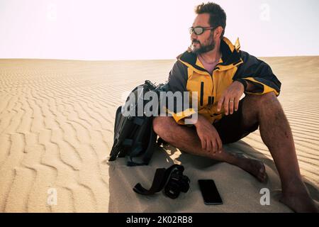 Der junge Erwachsene Mann erkundet Wüstensanddünen allein mit Rucksack und Fotokamera. Fotografie Landschaft Job-Aktivität mit bärtigen männlichen Menschen sitzen und ruhen nach barfuß auf Dünen Stockfoto