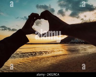 Pov des Paares tun Herz Geste Symbol mit Händen zusammen am Strand in Sommerurlaub Urlaub mit erstaunlichen Himmel und bunten Sonnenuntergang im Hintergrund. Menschen und Beziehungen Stockfoto