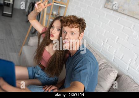 Junger Rotschopf-Mann, der Selfie mit Freundin macht, die Lippen ausschüttet und im Wohnzimmer ein Friedenszeichen zeigt, Stockbild Stockfoto