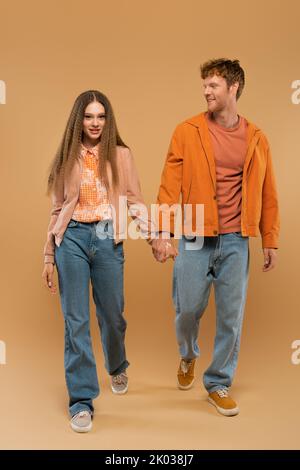 In voller Länge von glücklichen jungen Paar in herbstlichen Outfits und Gummischuhe halten Hände beim Gehen auf beige, Stock Bild Stockfoto