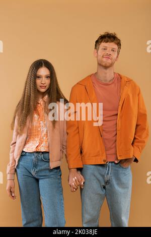 Glückliches junges Paar in herbstlichen Outfits, die Hände halten, während es isoliert auf beige steht, Stockbild Stockfoto