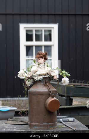 Milchkäntchen mit Puppe, weiße Rosen, dahinter Fassade eines Holzhauses, Marken-Halbinsel, Waterland, Noord-Holland, Niederlande Stockfoto