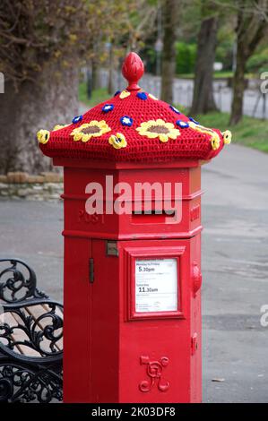 Skurriler, farbenfroher, handgestrickter Wollhut ziert in Solidarität mit der Ukraine eine alte rote viktorianische Säulenkiste mit gehäkelten Sonnenblumen. VEREINIGTES KÖNIGREICH. Stockfoto