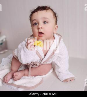 Ein kleiner Junge mit blonden Haaren und Augen in einem Bademantel sitzt auf einem Tisch. Stockfoto