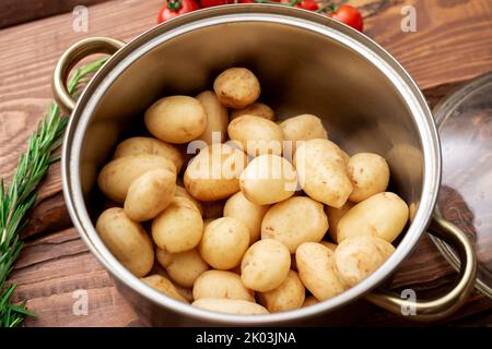 Rohe junge kleine Kartoffeln mit Rosmarin und Kirschtomaten in einem Topf auf braunem Holzboden. Kochen von gekochten Kartoffeln. Stockfoto