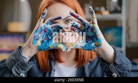 Nahaufnahme Kopfschuss rothaarige 20s lächelndes Mädchen in Brillen lächelnd toothy freundlich mit Liebe Malerin Frau Blick auf die Kamera hält schmutzig in Farben Stockfoto