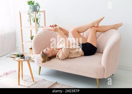 Die glückliche blonde Frau liegt auf dem Rücken auf einem beigen Sofa, die Beine sind hoch, schaut auf ein digitales Tablet Stockfoto