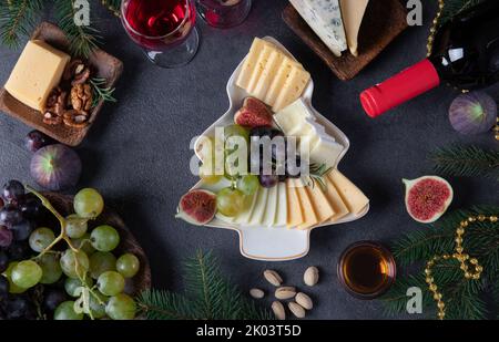 Verschiedene Käsesorten und Früchte, serviert auf einem Teller als Weihnachtsbaum, auf dunkelgrauem Hintergrund mit zwei Gläsern Wein. Silvesterparty-Snack. Draufsicht Stockfoto