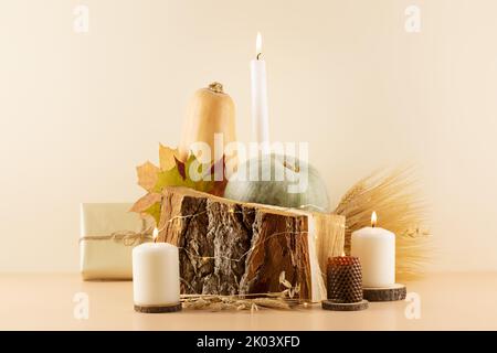 Erntedankfest Komposition mit Kürbissen, trockenen Herbstblättern, Bouquet von Roggen- und Weizenohren, Geschenken und Kerzen auf einem Holzpodium auf einem beigen Rücken Stockfoto