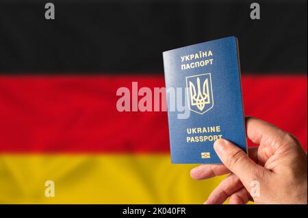 Ukrainischer biometrischer Pass in der Hand einer Person vor dem Hintergrund der Nationalflagge Deutschlands. Staatsbürgerschaft der Ukraine, Reisen nach Deutschland, Stockfoto