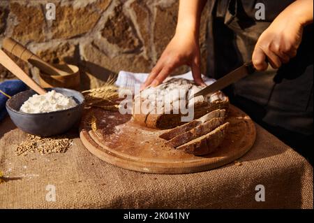 Baker schneidet einen Laib Roggenbrot auf Holzbrett. Weißes Mehl und Weizenspikeletts auf dem Tisch, bedeckt mit einer Quaste-Tischdecke Stockfoto