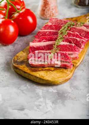 Frisches rohes Fleisch auf einem Holzbrett in Stücke geschnitten und Tomaten auf grauem Hintergrund. Gerichte aus Fleisch - Steaks, Steak, Suppe. Rezepte für Stockfoto