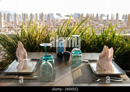 Leere Teller und Gläser, Stoffservietten auf dem Esstisch mit Blick auf das Meer und den Strand im Resort-Restaurant. Speicherplatz Kopieren. Stockfoto