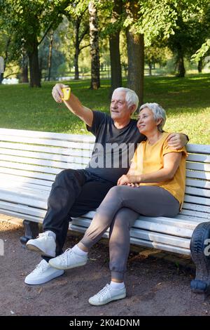 Glückliche ältere Ehepartner machen Selfie-Fotos Sitzen Sie auf der Bank im Sommerpark. Grauhaarige Paar Blick auf Handy-Bildschirm machen Videocall Outdoor-Nutzung modern t Stockfoto
