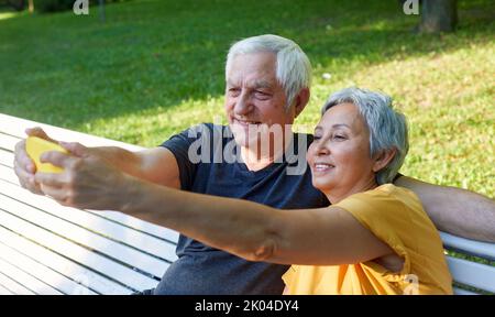 Glückliche ältere Ehepartner machen Selfie-Fotos Sitzen Sie auf der Bank im Sommerpark. Grauhaarige Paar Blick auf Handy-Bildschirm machen Videocall Outdoor-Nutzung modern t Stockfoto