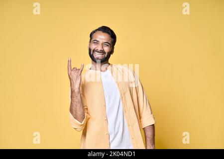 Glücklicher indischer Mann zeigt Rock n Roll Hörner Handgeste isoliert auf gelb. Stockfoto