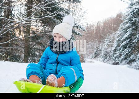 Junge sitzt auf dem Schlitten im winterverschneiten Land. Glückliches Kind Rodeln in der Natur. Kunststoffschlitten. Kind auf dem Schlitten im Wald. Stockfoto