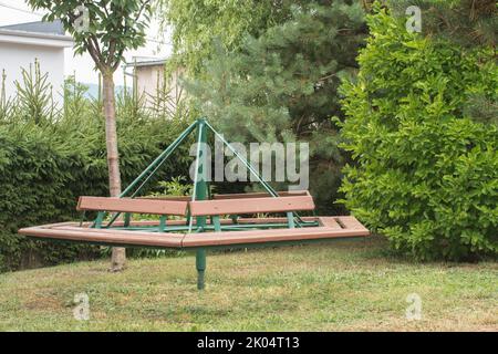 Retro-sozialistisches Kinderkarussell aus Holz und Eisen. Kinderroundabout im Hinterhof platziert. Umgeben von Gartenbelaubung. Hergestellt in der Tschechoslowakei. Stockfoto