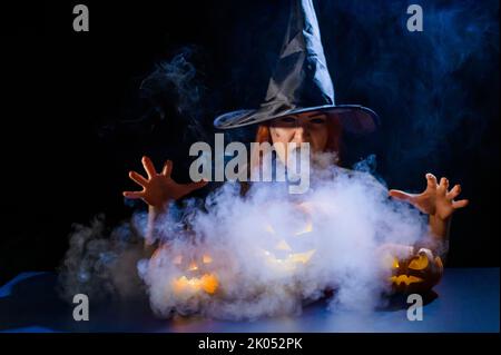 Eine ominöse Hexe im Hut zaubert über eine Jack-o-Laterne. Traditionelle halloween-Figuren. Mystischer Nebel kriecht über Kürbisse mit geschnitzten schrecklich Stockfoto