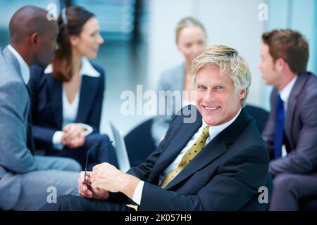Reifer Geschäftsmann lächelt selbstbewusst. Porträt eines selbstbewussten, reifen Geschäftsmanns, der lächelt, während er eine Brille mit Führungskräften im Hintergrund hält. Stockfoto