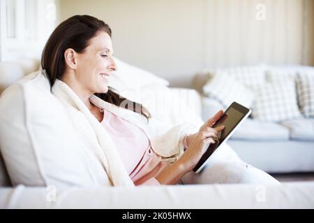 Lesen eines Romanromans auf ihrem E-Reader. Eine attraktive Frau, die ein digitales Tablet nutzt, während sie auf einem Sofa sitzt. Stockfoto