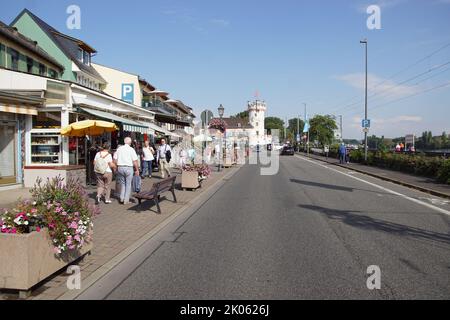Rheinstraße mit Restaurants und Geschäften und Touristen in der deutschen Stadt Rüdesheim entlang einer Eisenbahnstrecke und dem Rhein. Adlerturm aus dem 15.. Jahrhundert Stockfoto