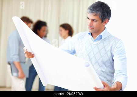 Geschäftsmann liest Blaupause. Porträt eines reifen Geschäftsmanns, der Entwürfe liest und im Hintergrund mit Kollegen diskutiert. Stockfoto