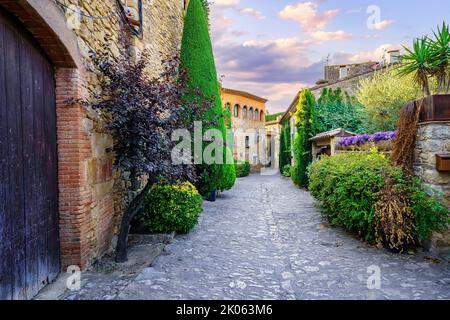 Alte Steinhäuser und Allee voller Pflanzen und Blumen bei Sonnenuntergang im Dorf Peratallada, Girona, Katalonien. Stockfoto