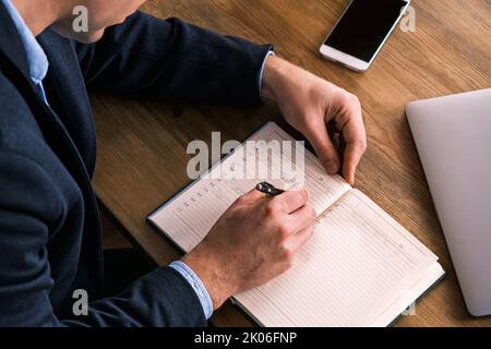 Junger seriöser Geschäftsmann schreibt etwas im Notebook, das mit dem liegenden Smartphone am Tisch sitzt. Mann trägt Brille füllt Notebook mit Stift, organisieren Stockfoto
