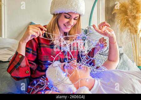 Weihnachten. Das junge Paar feiert Weihnachten zu Hause und liegt auf einem Bett und trägt weihnachtsmützen. Glücklicher Mann und Frau lächeln einander an. Der Mensch liegt auf Stockfoto