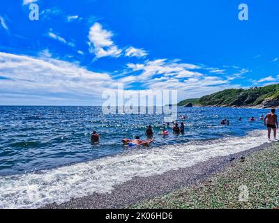 Wladiwostok, Russland - 25.08.2022: Am berühmten Steinstrand der Steklyannaya Bay schwimmen die Menschen. Strand aus natürlich poliertem strukturiertem Meerglas und Steinen an der Küste. Grünes, blau glänzendes Glas mit mehrfarbigen Meereskieseln aus der Nähe. Hochwertige Fotos Stockfoto