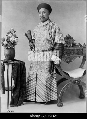 Wu-Ting-Fang, chinesischer Minister, in Washington, D.C., c1900. Dezember 6. Wu Tingfang, chinesischer Diplomat, Porträt in voller Länge, stehend, leicht nach links gerichtet, Einen Ventilator in der rechten Hand halten und die linke Hand auf dem Arm eines Stuhls ruhen; Tisch mit Vase und Blumen auf der linken Seite. Stockfoto