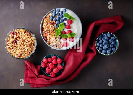 Joghurt mit Müsli und Beeren in einer Schüssel. Zusammensetzung mit gesundem Frühstück und Zutaten mit burgunderfarbener Serviette. Flach liegend Stockfoto