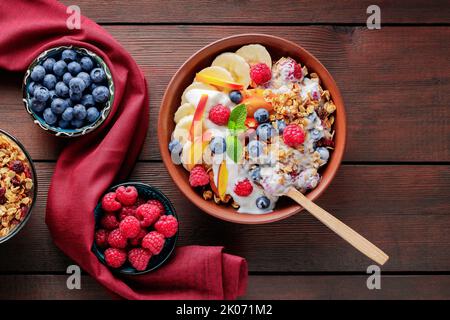 Griechischer Joghurt mit Müsli, Beeren und Früchten auf einem Holztisch. Gesundes Frühstück mit Müsli, Joghurt und Früchten auf einem Tonteller mit einem Holzlöffel Stockfoto