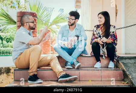 Drei junge Freunde, die auf der Treppe sitzen und sich unterhalten. Drei Teenager-Freunde, die sich auf der Treppe unterhalten, drei junge Leute, die auf der Straße sitzen Stockfoto